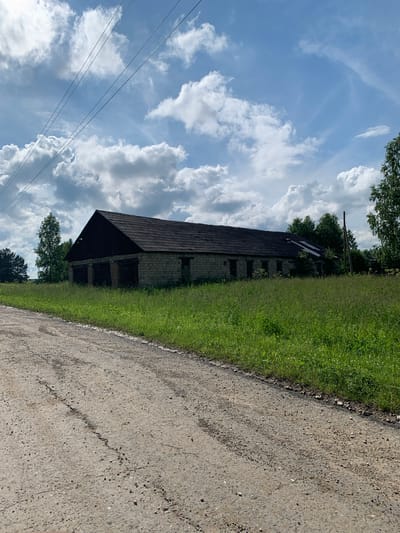 An exterior shot captures an abandoned, long, low-rise stone building with a dark, sloping roof. The building appears to be a barn or storage facility with several dark openings along its facade. It is surrounded by tall, green grass and foliage. A dirt road with tire tracks runs along the left foreground of the image. In the background, trees are visible, and a power line stretches diagonally across the upper portion of the frame. The sky is partly cloudy with dramatic white cumulus clouds against a blue backdrop. The scene is set in a rural area, potentially on the outskirts of Барсаи, Russia, during daylight.
