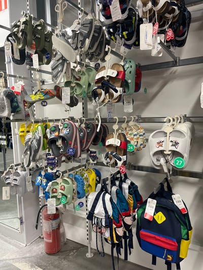 The image depicts a retail display of children's footwear and backpacks in a store. The items are hung on metal racks against a white wall. Several pairs of sandals, flip-flops, and clogs are visible, featuring various colors and cartoon character designs. The shoes are primarily organized by size, indicated by circular tags with numbers. A fire extinguisher is positioned on the floor in the lower left quadrant of the image. In the background, a reflection in a glass partition suggests the presence of more merchandise and store aisles. The lighting is artificial and bright, typical of an indoor retail environment. The overall scene is clean and orderly, with the merchandise neatly arranged for display and sale. The location of this scene is the LC Waikiki store at the Nikolsky mall in Kharkiv, Ukraine.