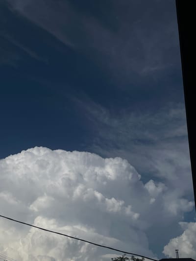 The media file shows a view of the sky from below, looking upwards. The sky is divided into two distinct sections: a deep blue, almost black, portion on the left, and a bright white, cloudy portion on the right. Wispy, thin clouds are visible in the darker section, while the right side is dominated by large, billowing cumulus clouds. A single dark line, likely a power line, runs diagonally across the right side of the frame. There are also some small, pinkish-purple dots visible in the sky, which could be distant balloons or other small airborne objects. The top of the frame is partially obscured by a dark, horizontal band, possibly the edge of a building or structure. The overall impression is of a bright, possibly sunny day with some dramatic cloud formations. The time of day appears to be daytime, given the brightness of the clouds and the blue sky. There are no visible people or text in the image. The location context provided is Novopokrovskaya, Russia.