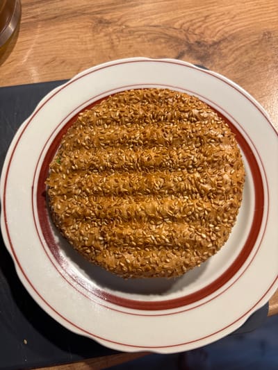 A close-up, overhead shot shows a round, toasted bread roll covered in sesame seeds, resting on a white plate with a red rim. The bread has grill marks. The plate is situated on a wooden surface, with a dark surface to the left. A portion of a glass container with a golden liquid is visible in the upper left corner. This image was taken in Kemerovo, Russia.