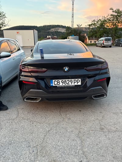 A black BMW 8 Series coupe is parked outdoors on an asphalt lot. The car has a Bulgarian license plate reading "CB 9829 PP". Below the license plate, the text "REAPERS KOSTINBROD" is visible. A blue and silver can sits on the trunk of the car. To the left of the BMW, a portion of a silver car is visible, reflecting a sunset sky. In the background, a white portable toilet structure with "WC" signage is present, along with trees, a hill with mining activity, and a cell tower. The sky displays the colors of a sunset. The ground is a rough asphalt surface with some cigarette butts scattered on it.