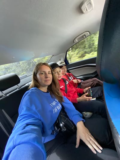 A selfie taken from inside a vehicle shows a woman in the front seat and two young boys in the back. The woman, with long brown hair and a blue sweatshirt, is looking at the camera. The boy to her right, wearing a red hoodie and a black baseball cap, is also looking at the camera. The other boy, further back and to the right, is wearing a red hoodie and a white baseball cap. The interior of the car is visible, as is the blurred greenery of trees and foliage passing by outside the window, suggesting the vehicle is in motion. The image does not provide any specific details that would pinpoint a location within the city of Niva, Russia.
