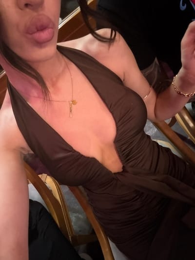 The image displays an individual, likely female, from the upper torso upwards, angled slightly to the right. The lower portion of her face is visible, with lips pursed in a "kissing" pose. She wears a dark brown, deeply V-necked dress made of ruched fabric. A delicate gold-colored necklace with two small pendants (one disc-shaped, one bar-shaped) rests on her chest. Her left wrist bears a thin gold-colored bangle, and her right wrist has a wider gold-colored chain-link bracelet. The skin on her chest and shoulders exhibits pinkish light reflections and subtle glitter. The background is an indistinct, dimly lit indoor environment, with parts of a wooden chair visible, suggesting the individual is seated. No visual information within the image provides details to identify a specific geographical location, such as Debrashtitsa, Bulgaria.