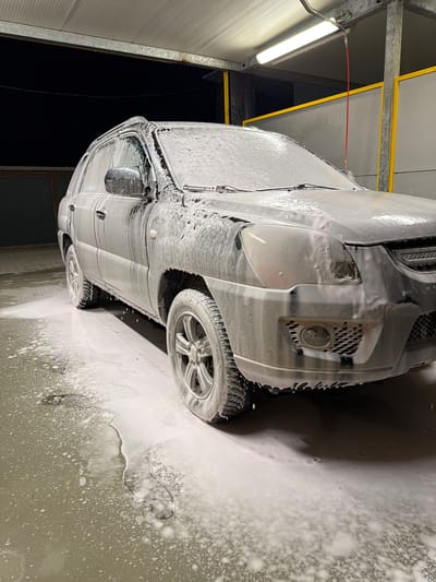 A silver SUV, covered in white foam, is being washed at an outdoor, covered car wash in Pernik, Bulgaria. The vehicle is positioned at an angle, with the front driver's side wheel and part of the front bumper visible. The ground around the car is wet and covered in foam. The car wash structure, with a metal roof and yellow and gray walls, is visible in the background. A bright fluorescent light illuminates the scene. The time appears to be night, indicated by the dark background. There are no people visible in the image.