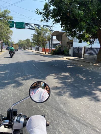 The image depicts a street scene in Madiun, Indonesia, viewed from the perspective of a person riding a motorcycle. In the foreground, a gloved hand grips the motorcycle's handlebar, and a portion of the rider's torso is visible in the side mirror, wearing a white shirt with red trim. The road surface is asphalt, with visible tire tracks and scattered debris. Sunlight casts tree shadows across the road.

Along the side of the road, there are several buildings, including a white structure with a black metal fence and potted plants. Further down the street, trees line the roadside, and a green traffic sign with white text is visible, indicating directions to "ROGO," "KOTA," "RSU SOEDONO," and "STASIUN KA." A yellow truck and other motorcycles are present on the road, suggesting moderate traffic. The sky is clear and blue.