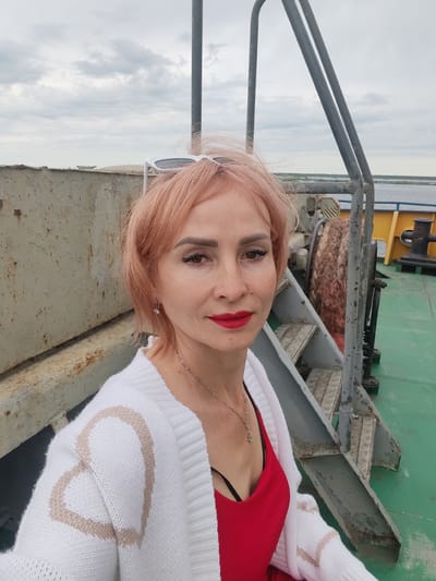 A close-up, waist-up selfie of a woman with short, light pink hair and bright red lipstick, wearing a white cardigan with a heart pattern over a red top. She is on what appears to be the deck of a boat or ship, with a metal railing and staircase visible behind her. The environment is overcast with a cloudy sky. The location is identified as Bereg, Russia.