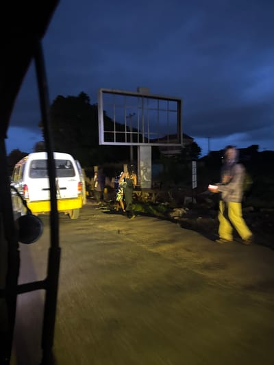 The image depicts a street scene at dusk in Enugu, Nigeria, characterized by a dark, cloudy sky and the presence of street lighting and artificial light sources. In the foreground, a portion of a vehicle, likely a three-wheeled auto-rickshaw or similar transport, is visible on the left, with dark metal bars framing the view.
A white van with a yellow lower body is parked on the right side of the road, facing away from the camera. Several people are gathered near the van. One individual, wearing a grey hooded jacket and yellow pants, is walking from right to left across the road, holding a glowing rectangular object, possibly a phone or a small light. Behind the van, a group of people are standing and interacting. One person in the group is wearing a black dress, and another is in a blue and white patterned outfit.
Further in the background, a large, empty billboard structure stands prominently, with a grid-like frame. The surrounding environment shows signs of urban infrastructure, with some vegetation and potentially other buildings or structures obscured by the low light conditions. The road surface appears to be paved and wet, reflecting the available light. The overall impression is a typical evening scene in a Nigerian city, with a sense of movement and activity despite the fading light.