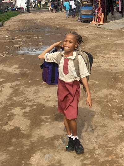 A young girl, likely a student, is the central figure in this daytime outdoor shot taken on a street in Ijegun, Nigeria. She stands on a rough, unpaved or poorly paved road that appears muddy or wet in patches, with varied shades of brown, tan, and grey.

The girl has dark skin and her hair is styled in braids. She wears a school uniform comprising a light-colored collared shirt with maroon accents and a tie-like embellishment, and a maroon knee-length skirt. A large backpack, a combination of tan, blue, and purple, is strapped to her back. She completes her attire with white socks and black athletic shoes that feature a subtle light green detail near the toes. Her face is turned towards the camera, beaming with a wide, joyful smile, her eyes squinted, possibly due to bright sunlight.

In the background, to the left of the girl, is a bustling street scene with several market stalls or temporary shops displaying various fabrics and goods. People are visible near these stalls, engaged in daily activities; one person wears a light blue shirt, another a white top, and others are partially obscured. Further down the road, several motorcycles or scooters can be seen parked. A narrow strip of green grass lines the far left edge of the scene.

The bright lighting suggests it is daytime. The girl's cheerful expression and uniform indicate she is either on her way to or from school, bringing a sense of youthful energy to the busy market environment. There is no visible text in the image.