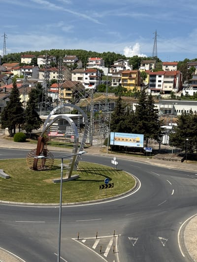 This is an outdoor, eye-level photograph depicting a cityscape with a traffic roundabout in Bragança, Portugal.
The roundabout has a grass-covered center island with road signs. A modern art installation made of metal curves is located near the left side of the island. A billboard with the word "VERDADE" (meaning "truth" in Portuguese) is visible on the side of the road. A second sign is partly visible, displaying election slogans and other political messaging.
In the background, there are residential buildings of various colors and architectural styles, interspersed with trees. Electrical pylons and power lines can be seen crisscrossing the sky and the landscape between the buildings. The sky is blue with sparse clouds. The photo is taken during daylight, with good natural lighting.
