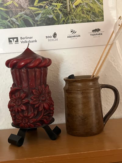 A close-up, eye-level shot depicts a still life on a wooden shelf against a white wall, with a poster featuring bamboo foliage and logos of Berlin-based establishments visible in the background. On the left, a dark red, ornate candle shaped like a cupcake with floral carvings sits on a black metal stand. To its right, a rustic brown ceramic jug holds a dark object and two light brown wooden sticks. The jug is positioned slightly in front of the candle. The poster, predominantly showcasing green bamboo leaves, also displays logos for Berliner Volksbank, Zoo Berlin, Aquarium Berlin, and Tierpark Berlin, with text indicating "Großer Panda im Zoo Berlin | Foto: Karl Bröseke." The lighting is even and diffused, illuminating the objects clearly.