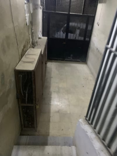 An indoor space with a low ceiling featuring exposed pipes. A large metal cage-like structure sits on the floor, connected to a platform. A second, similar structure rests nearby. A metal door is visible to the left, and the walls appear to be concrete or a similar material. The lighting is dim, and the overall scene suggests an industrial or utility area.