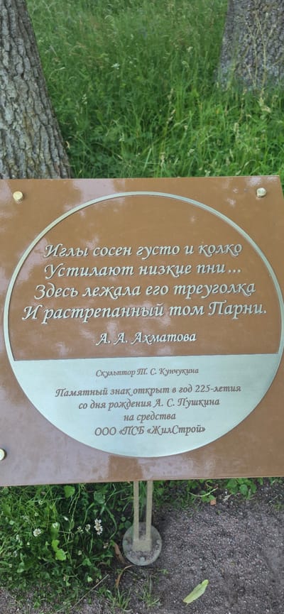 The image shows a memorial plaque with text inscribed on it, placed outdoors near a tree and grass. The plaque is round and has a metallic sheen, with text etched in a lighter color. The text appears to be a poem or excerpt of literature, followed by attribution to A. A. Akhmatova. Below this, there is information about the sculptor, the year of the memorial's opening, and the patron. The background consists of lush green grass, a tree trunk on the left, and a gravel path at the bottom. The overall impression is that of a historical or literary monument located in a park or garden setting. The specific location is stated on the plaque as being in the city of Pushkin, Russia, commemorating the 225th anniversary of A. S. Pushkin's birth.