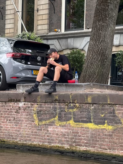 A person wearing a black t-shirt, black shorts, black sneakers, black socks, and a black baseball cap is sitting on the trunk of a grey Volkswagen car, eating a sandwich. The car is parked next to a brick wall that has yellow spray paint graffiti on it. The person's legs are bent, and they are holding the sandwich up to their mouth with both hands. The scene appears to be outdoors, possibly on a street in Amsterdam, Netherlands, given the canals often found in the city. The time of day is likely daytime, as indicated by the natural light. There is a water bottle and a red object on the trunk of the car. The overall mood appears casual and relaxed, with the person focused on their meal. The license plate of the car is visible and reads "NL GXV".