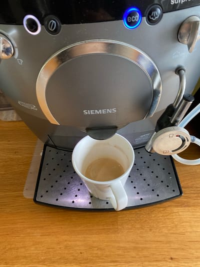 A Siemens espresso machine sits on a wooden surface. The machine has a cup on its drip tray. The "eco" button is illuminated blue. The dial for milk frothing is visible. The scene is possibly indoors. Location cannot be determined without further information.