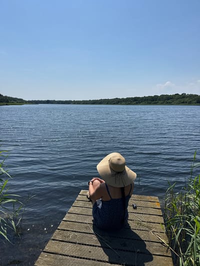 A person wearing a wide-brimmed straw hat and a dark blue dress with white polka dots sits on a wooden pier next to a body of water. The person is facing away from the camera, looking out at the water. The pier is made of weathered wooden planks and is partially covered by a wire mesh. Green reeds grow along the edge of the water. The water is rippled and reflects the sunlight. In the distance, a tree line forms the far bank of the lake. The sky is a clear, bright blue with a few scattered clouds. The scene suggests a peaceful, sunny day. The image appears to have been taken in the afternoon, given the quality of the light.