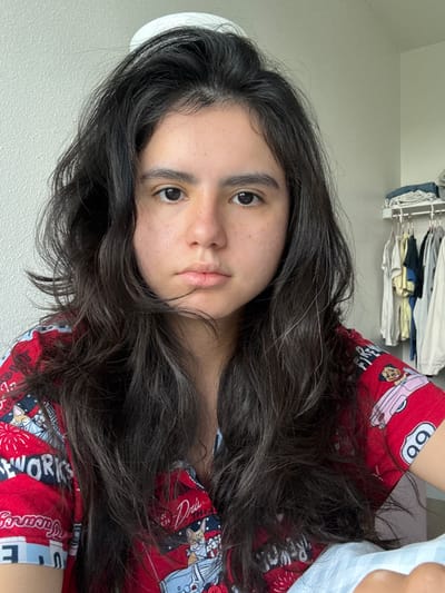 The image presents a close-up, portrait-oriented shot of a young person, likely female, positioned horizontally with their head resting on a surface, looking directly at the camera. The subject has long, dark, wavy hair that flows around their face and extends across their chest. Their complexion is light to medium with noticeable freckles on their cheeks, and they have dark eyes and full lips, maintaining a neutral and calm expression. Individual strands of white or grey hair are visible amidst the dark hair.
The person is wearing a red patterned top featuring various cartoon graphics. Distinct designs include a pink car with a character and the number "66" enclosed in a white circle, as well as illustrations resembling fireworks. Visible text fragments on the shirt include "FIREWORKS," "Drie," and "Fancy."
The background is a light-colored wall, indicating an indoor setting, likely a home environment. Above the subject, a clothing rack with various garments, such as t-shirts and folded fabrics, is visible, suggesting a personal space like a bedroom. The lighting is soft and even, consistent with natural daylight indoors. The overall atmosphere conveyed is relaxed and quiet.