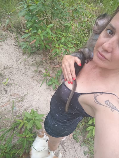 A woman is holding a young monkey on her shoulder. The woman has fair skin, dark hair pulled back, and is wearing a dark-colored sleeveless top. She has red nail polish and a tattoo on her upper arm. The monkey is small, with dark fur and a long tail. Another monkey is partially visible behind the woman. In the foreground, at the woman's feet, is a large white and orange cat lying in the grass. The background consists of sandy ground with patches of green vegetation and small plants. The location is Ban Tai, Thailand.