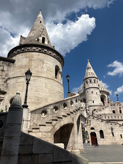 The image captures a detail of Fisherman's Bastion in Budapest, Hungary, on a bright, partly cloudy day. The perspective is from below, looking up at two of the iconic conical towers. The architecture is predominantly light-colored stone, with intricate carvings and decorative elements. One tower in the foreground is larger and closer, showcasing a circular base with arched windows and a sharply pointed conical roof topped with a finial. A black lamppost with an ornate design stands in front of this tower.
Behind the foreground tower, a second, taller tower is visible, also conical and adorned with similar stonework, featuring multiple levels of windows and decorative bands. The broader structure of the bastion is visible behind this tower, with a facade made of rectangular stone blocks, arched openings, and small windows. Some circular cutouts are visible on the bastion's walls, adding to its unique design.
A bright blue sky with scattered white clouds forms the backdrop. The lighting suggests it is daytime, likely late morning or early afternoon, given the strong shadows cast by the architectural elements. The scene conveys a sense of grandeur and historical significance. While no specific event is depicted, the presence of the architecture and the sky suggests a typical day at this tourist landmark. There is no visible text in the image.