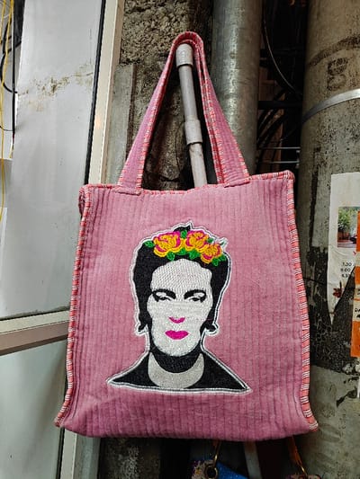 A pink corduroy tote bag, featuring an embroidered image of Frida Kahlo's face with a yellow and orange floral crown and fuchsia lips, hangs in an urban setting within Kochi, India. The bag, characterized by vertical ribbed texture and striped pink and white trim on its handles, is suspended from a grey pipe. The background consists of a stained, light grey concrete wall. To the left, a white vertical building panel or frame is partially visible with yellow and black electrical wires. To the right, another vertical grey pipe and a weathered, partially obscured white paper poster with black text, including timings (e.g., "7:30 am", "3.00 pm", "4.30 pm") and Malayalam script, are visible against the wall. No individuals are present in the frame.