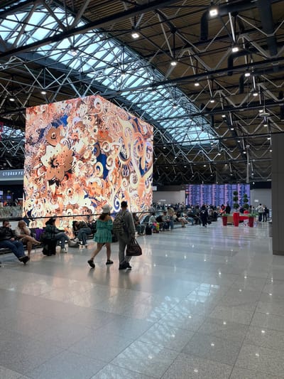 The image depicts the interior of a modern airport terminal, likely located in or near Перепечино, Russia, given the visible signage. The space is vast and open, with a high, industrial-style ceiling featuring a complex network of metal beams and a translucent roof that allows natural light to filter in. Large, bright LED screens dominate the scene. One massive cube-shaped screen displays an intricate, abstract floral pattern in warm orange, gold, and blue tones. In the background, another large screen shows a departure or arrival board with rows of text and numbers.

The floor is polished and reflective, made of large gray tiles. Several groups of people are seated on benches, waiting, with various pieces of luggage around them. In the foreground, two individuals are walking away from the camera. One is a woman in a short, light blue dress and a white bucket hat, and the other is a man carrying a dark brown bag. The overall atmosphere is that of a busy, well-lit transportation hub.