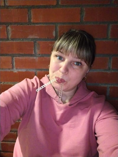A woman is captured in a close-up portrait, looking directly at the camera with a playful or curious expression. She has light brown or dark blonde hair with bangs covering her forehead and prominent blue-green eyes. Her mouth is pursed around a translucent, segmented plastic object that features small circular holes along its length. She is wearing a pink hooded sweatshirt and small golden hoop earrings. The background consists of a red brick wall with dark mortar, suggesting an indoor setting. There is no visible text in the image. The location is contextualized as Rusanovo, Russia.