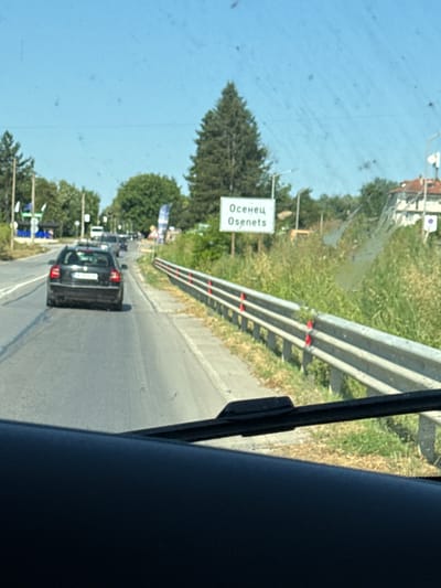 The image shows a road scene in Osenets, Bulgaria, viewed from inside a vehicle. A black car is driving in the right lane on a paved road. There is a white sign with Cyrillic and Latin text that reads "Осенец Osenets". The sign appears to be a town limit sign. A metal guardrail runs along the right side of the road, separating it from a grassy area. Trees and buildings are visible in the background. The sky is clear and blue, suggesting a sunny day.