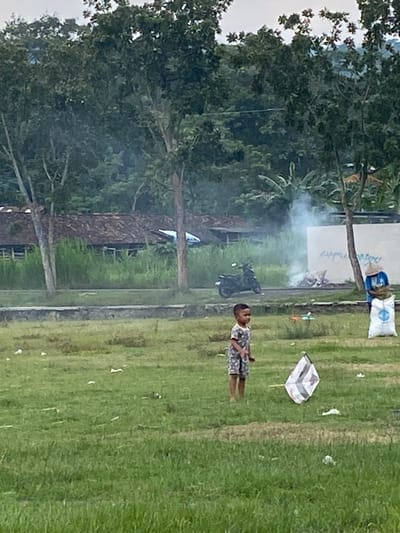 The scene takes place in a grassy field in Kecamatan Piyungan, Indonesia, located at coordinates -7.828 latitude and 110.496 longitude. A young boy, wearing a patterned shirt and shorts, stands in the field holding a white kite with pink and black stripes. Smoke drifts in the air, possibly from a recent fire, and a motorbike is parked nearby. A person wearing a blue shirt and a hat stands in the background, holding a large white bag. Several pieces of trash, including white plastic bags, are scattered across the field. In the background, a row of buildings with a tiled roof can be seen, surrounded by trees. There are trees on either side of the field, and a fence runs across the foreground. The ground is covered in green grass. There is no other visible activity in the image.