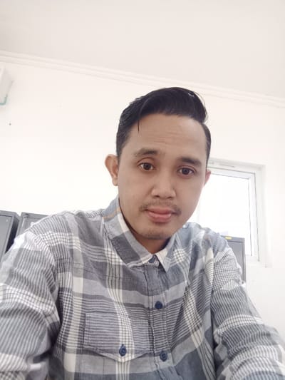A man poses in a gray and white plaid shirt, buttoned up to the collar, with a small pocket visible on the left. He has dark hair styled back and a faint mustache. The background is a bright white, with a window and filing cabinets partially visible. The man's expression is neutral, and the shot is taken from a slightly high angle.