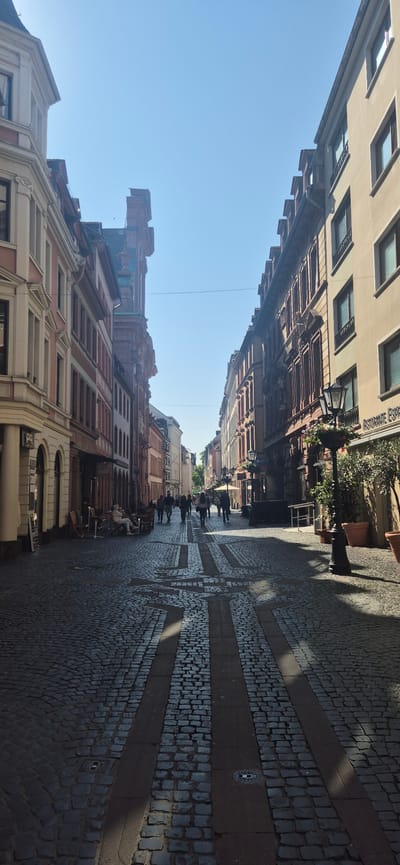 The image depicts a narrow cobblestone street lined with historic buildings in Mainz, Germany. The street is a pedestrian zone with several people walking and seated at outdoor cafes. On the left, there are cream-colored buildings with red accents and arched doorways. On the right, there are buildings with reddish-brown facades, featuring numerous windows and decorative elements. A black lamppost with hanging flowers stands on the right side of the street, near a restaurant with a sign that partially reads "Ristorante Espace". The cobblestone pavement has a pattern of darker stones forming geometric shapes. The sky is clear and blue, indicating a sunny day. The perspective of the image is looking down the street, creating a sense of depth.