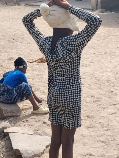The image depicts a dusty outdoor scene in Gwagwarwa, Nigeria, featuring two individuals. In the foreground, a person, viewed from behind, stands with both hands raised to the top of their head, appearing to adjust a cream-colored knitted beanie with a pompom. This individual wears a long-sleeved dress or tunic with a black and white houndstooth pattern. Dark-skinned legs are visible below the garment's hem. To the left and slightly behind, a second individual sits on a light-colored rock or concrete block. This person wears a blue long-sleeved top, patterned blue and white lower garment, and a dark head covering with a white band. One foot, clad in a light yellow Crocs-style sandal, is visible. The ground is unpaved, consisting of light brown earth and sand with scattered light-colored debris. In the distant background, blurred structures with light-colored walls are visible.