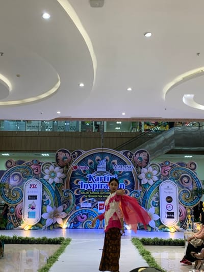 The image shows a fashion show taking place in the Malang Town Square in Malang, Indonesia, located at coordinates -7.957 latitude and 112.619 longitude. A young woman wearing a traditional Indonesian outfit is walking down a runway, smiling and waving her hands. The background is a large colorful stage with the words "Kartika Inspirasi" written on it, along with the logo of the Malang Town Square and several sponsors. The runway is lined with green plants. The scene takes place inside a shopping mall, with a white ceiling and an escalator visible in the background. Some members of the audience are seated in the foreground.