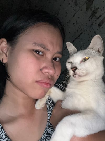 A selfie taken at night shows a young woman holding a white cat in East Jakarta, Indonesia. The woman, positioned on the left side of the frame, has her face turned towards the camera with a displeased expression, her mouth slightly pursed. She has dark hair pulled back and is wearing a hoop earring in her left ear. The cat, on the right, is white with light grey markings, and appears to be winking, with its head tilted. The background is dark and indistinct, suggesting an indoor setting.