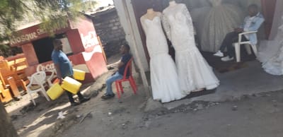 A streetside scene features a Coca-Cola-branded kiosk with a man carrying yellow jerrycans. Another man sits on a red plastic chair. A bridal shop is adjacent, displaying two white wedding dresses on mannequins. A third man sits on a white chair within the shop's doorway, partially obscured by more wedding dresses. The area appears dusty and unpaved.