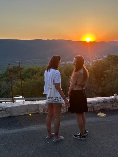 Two individuals, a woman with dark hair wearing a white t-shirt and denim shorts, and another woman with long auburn hair wearing a black bikini top and a black skirt, are standing on a paved surface near a stone wall. The woman with dark hair is holding a white tote bag with text that reads "BITCOIN maximalists." Both are facing each other and appear to be in conversation. The background shows a hilly landscape with trees and a village in the distance, illuminated by a vibrant sunset. The sun is setting behind the hills, casting a warm orange glow across the sky. The location is identified as Svinjišta, Montenegro.