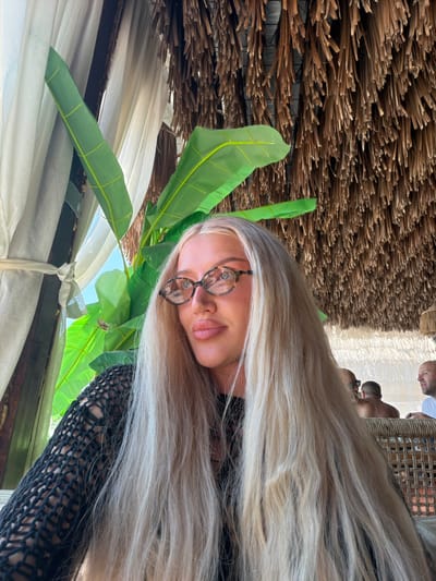 In Drobnići, Montenegro, a young woman with long, blonde hair and glasses is seated at a table, with her left side facing the camera. She is wearing a black, mesh top. Behind her, large green leaves from a tropical plant are visible, along with a thatched roof ceiling and sheer curtains. In the background, two men are seated, one of whom is looking towards the camera. The setting appears to be an outdoor cafe or restaurant.