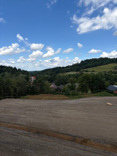 The image depicts an outdoor scene in the rural outskirts of Stróża, Poland, under a bright blue sky dotted with white clouds. The foreground is a construction site with a large, flat area of compacted dirt, marked by tire tracks. A yellow caution tape runs along the edge of this area. Beyond the construction site, the landscape transitions into rolling green hills covered with dense forests. A few scattered houses with red and white roofs are visible nestled among the trees. The overall atmosphere suggests a sunny day in a peaceful, semi-rural environment that is undergoing development.