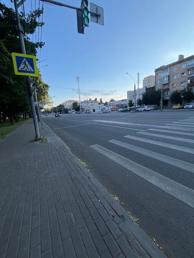 The image depicts a street scene in Voronezh, Russia. The viewpoint is low, looking up at a clear blue sky and across a wide road. A pedestrian crossing, marked by white stripes on the asphalt, dominates the right side of the frame. The sidewalk, paved with dark grey bricks, runs along the road.

On the left side of the road, a traffic light with green lights is visible, along with a blue and yellow diamond-shaped road sign depicting a pedestrian walking. Behind the sign, trees and utility poles with wires can be seen. Further down the street, apartment buildings with multiple stories line the road, some with balconies. Cars are driving on the road, and there are some small kiosks or stalls on the sidewalk.

The image appears to have been taken during the daytime, likely in the late afternoon or early evening given the angle of the sun and the shadows. The weather seems to be clear and sunny. The overall impression is of an urban environment on a typical day. There is some text visible on one of the kiosks, but it is too blurry to read.