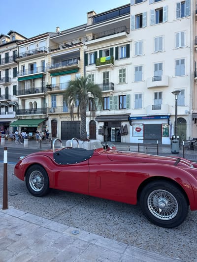 A red convertible classic car is parked on a cobblestone street in Cannes, France, with a row of buildings lining the background. The buildings feature multiple stories with balconies and shuttered windows. A palm tree stands near the car. Several people are walking on the sidewalk, and some are seated at outdoor tables in front of shops and restaurants. The sky is clear and blue.