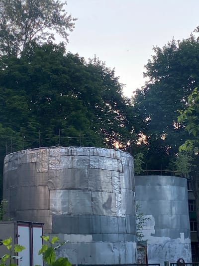 The image depicts an outdoor scene in Podolsk, Russia, with a focus on two large cylindrical structures covered in silver sheeting, possibly water tanks or industrial equipment undergoing repair or construction. The sheeting is patched in places, suggesting ongoing work or a rustic aesthetic. Lush green trees surround the structures, with some sunlight filtering through the leaves, indicating it is daytime, likely late afternoon due to the soft light. The sky is overcast and bright, typical of a temperate climate. A few small birds are visible in the sky. The overall impression is one of an industrial or utility area set against a natural backdrop. There is no visible text in the image.
