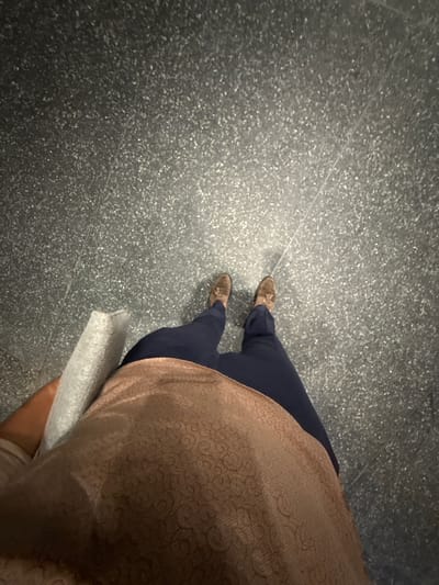 The image is a high-angle, full-body shot of a person standing on a speckled grey floor, likely indoors. The person is wearing dark navy pants and a light brown, textured top. They are also wearing brown suede loafers with tassels. In their left hand, they are holding a light grey, rectangular object wrapped in what appears to be a protective material. The floor has visible lines, suggesting it could be tiled or a sectioned surface. The lighting appears to be overhead, casting faint shadows. There are no discernible landmarks or context to place this scene within Vancouver, Canada.