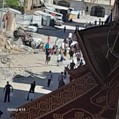 The image, rotated 90 degrees clockwise, captures an outdoor scene from an elevated perspective in Khan Yunis, Palestinian Territories. A large, dark brown fabric or canopy with intricate geometric and curvilinear patterns, likely providing shade, dominates the top and right portions of the frame.
Beneath this overhead covering, a street or open area is filled with numerous people. Many individuals are seen lying down or seated on white sheets and blankets spread on the ground, suggesting they are resting or sleeping in a temporary encampment. Their attire is casual and varied. Other figures are standing, casting distinct, elongated shadows indicative of bright sunlight.
Various objects are scattered across the ground, including personal belongings, clothing, and what appears to be debris, further emphasizing the makeshift nature of the setting. In the background and along the left side of the image, urban structures and buildings are visible, some appearing worn or damaged.
The clear, bright lighting indicates it is daytime. The overall scene portrays a situation of displacement or temporary outdoor living. In the bottom right corner, the text "Galaxy A14" is visible, indicating the device used to capture the photo.