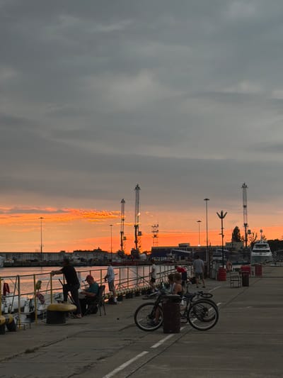 This image captures a serene sunset scene at a pier in Sirius, Russia. The sky is painted with vibrant hues of orange and yellow, transitioning into a muted gray as it ascends. In the foreground, a concrete pier stretches out, with several people present. A man in a black t-shirt stands leaning against a railing, looking out towards the water. Nearby, another man sits in a chair, engrossed in a book, while a young person stands next to him. Further down the pier, a group of people are gathered, some appearing to be having a conversation. Two bicycles are parked by the pier, one of them with a child seated on the back. The background reveals a harbor with various boats docked and industrial cranes reaching towards the sky. The overall atmosphere is one of peaceful evening leisure by the sea.