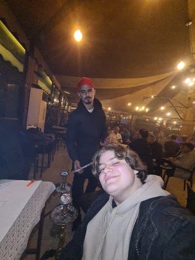 Two individuals are centrally framed. The person on the right has blond, curly hair and wears a light-colored hoodie under a black jacket. The person on the left sports a red beanie and a black jacket. Both are seemingly engaged in smoking a hookah, seen positioned between them. They are indoors, likely in a dimly lit lounge or restaurant, indicated by string lights overhead and a crowd in the background. The photo is taken at a tilted angle, adding an informal, casual vibe.