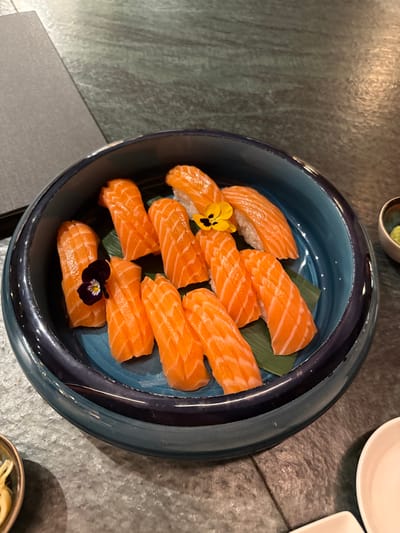 The image displays a culinary presentation of salmon nigiri sushi in an upscale restaurant setting within Bordeaux, France. The central subject is a deep, round ceramic bowl, predominantly dark blue with a lighter blue interior, filled with approximately twelve pieces of salmon nigiri. Each piece features a vibrant orange slice of salmon draped over white rice, with some salmon exhibiting fine scoring patterns. The nigiri are arranged on two green, rectangular decorative leaves at the bottom of the bowl. Two pieces of sushi are garnished with small edible flowers: one with a purple and yellow pansy-like bloom, and another with a solid yellow flower featuring a dark center. The bowl rests on a dark, mottled grey-green tabletop, possibly a stone or textured laminate. Partially visible on the table are several other items: a dark, rectangular object to the upper left (likely a menu or check holder), a small dark-rimmed bowl with a pale green substance (presumably wasabi) to the right, a small white ceramic bowl to the bottom right, and a small, light-colored bowl containing pale yellow, thinly sliced ingredients (likely pickled ginger) to the bottom left. No individuals or specific actions are depicted.
