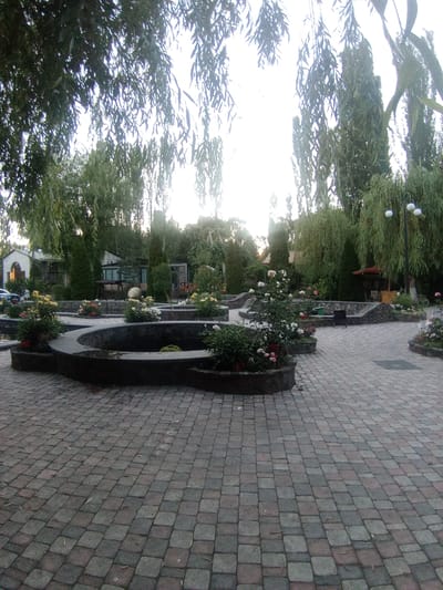 This is an outdoor, eye-level, wide-angle shot of a paved courtyard area with several water features and surrounding greenery. In the foreground, a curved, dark stone wall encloses a shallow water feature. Beyond this, a larger, more elaborate water feature is partially visible, with stone borders and some plants. The courtyard itself is paved with interlocking bricks in shades of gray and reddish-brown.
Several beds of flowering plants, primarily roses in pink and white, are scattered throughout the courtyard and around the water features. Tall, slender trees, some with drooping branches reminiscent of weeping willows, line the background, creating a lush, green environment. In the mid-ground, behind the water features, are several small buildings, likely part of a guesthouse or restaurant, with stucco exteriors and darker roofs. One building has a porch with a swing-like seat. Further in the background, more trees and vegetation are visible. A lamppost with three white globe lights stands to the right of the frame. A bicycle is leaning against a wall in the far right background.
The lighting suggests it is late afternoon or early evening, with soft light and long shadows. The sky is bright and overcast. The overall atmosphere is serene and tranquil, evoking a sense of a peaceful garden retreat. There is no visible text in the image. The location cues (buildings, vegetation style) align with the provided context of Katnaghbyur, Armenia. No people are visible in the scene.
