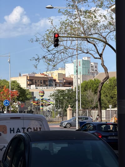 The image depicts a street scene in Castelló de la Plana, Spain. In the foreground, the top of a dark-colored car is visible, with a white van to its left bearing the "HATCHI" logo. Behind the van, a silver car and a dark blue car are stopped at a traffic light, which is red. Further in the background, buildings of varying heights and architectural styles line the street. A distinctive green building with the word "CASTELLON" is prominently visible. The street is lined with trees, one of which has red berries. A large, colorful abstract sculpture is positioned in the middle ground, adding an artistic element to the urban landscape. The sky is blue with scattered white clouds, suggesting fair weather. Several pedestrians are present in the scene, including a woman with a colorful bag walking on the right side of the image. A roundabout sign is visible on the left.