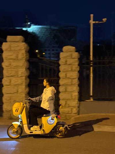 A person is shown standing next to a yellow electric scooter in the city of Zhongtun, China. The person is wearing a light-colored hooded jacket with blue sleeves, dark pants, and glasses. The scooter is parked on a concrete surface with a barrier of repeating rectangular blocks behind it. In the background, a street light stands next to buildings reflected in a dark body of water.