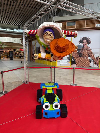 This image depicts an indoor exhibition within a shopping mall in Mont-Saint-Martin, France. A large LEGO sculpture of Buzz Lightyear, primarily white, green, and purple, is suspended from an overhead metal truss structure, appearing mid-flight and holding an orange LEGO cowboy hat in its right hand. Directly beneath, on a red carpet, is a smaller LEGO sculpture of an RC car, colored blue and lime green with black wheels and two prominent white and black 'eyes'. The red carpet is demarcated by silver stanchions connected by red ropes. The background features a large graphic advertisement of a woman in a brown polka-dot dress, wooden wall paneling, and the tiled floor and shopfronts of the mall's interior.