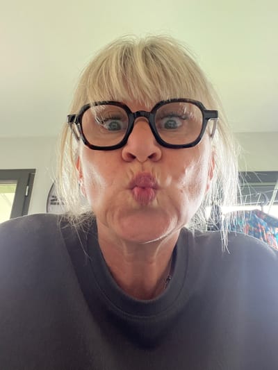 A woman with short blonde hair is centered in the image, wearing black-rimmed glasses and a dark-colored shirt. Her lips are pursed in a kissing gesture. The background includes a white wall on the left side and, behind her head, a clothes-drying rack. A dark-framed door with a visible green exterior is in the lower center of the image. A clock is present in the background as well. There are no other identifiable people, subjects, objects, actions, interactions, or environmental details to pinpoint the location in Chambœuf, France.