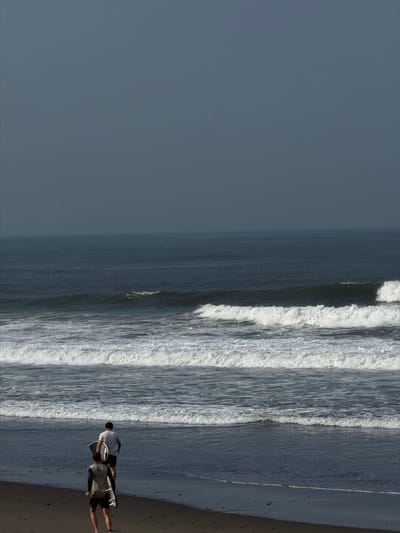 Two males are observed from behind, walking barefoot towards the ocean on a dark sandy beach in Belalang, Indonesia. The individual in the foreground wears a light sleeveless top over a dark long-sleeved garment and dark shorts, carrying a light-colored surfboard under his left arm. The individual positioned further forward wears a white short-sleeved t-shirt and dark shorts, holding a dark bodyboard with a white underside under his right arm. The ocean features multiple parallel lines of white-capped waves breaking towards the shore. The sea extends to the distant horizon under an overcast, dark grey sky.